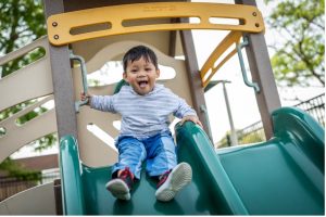 Toddler smiling and sliding down a blue plastic slide at an outdoor playground